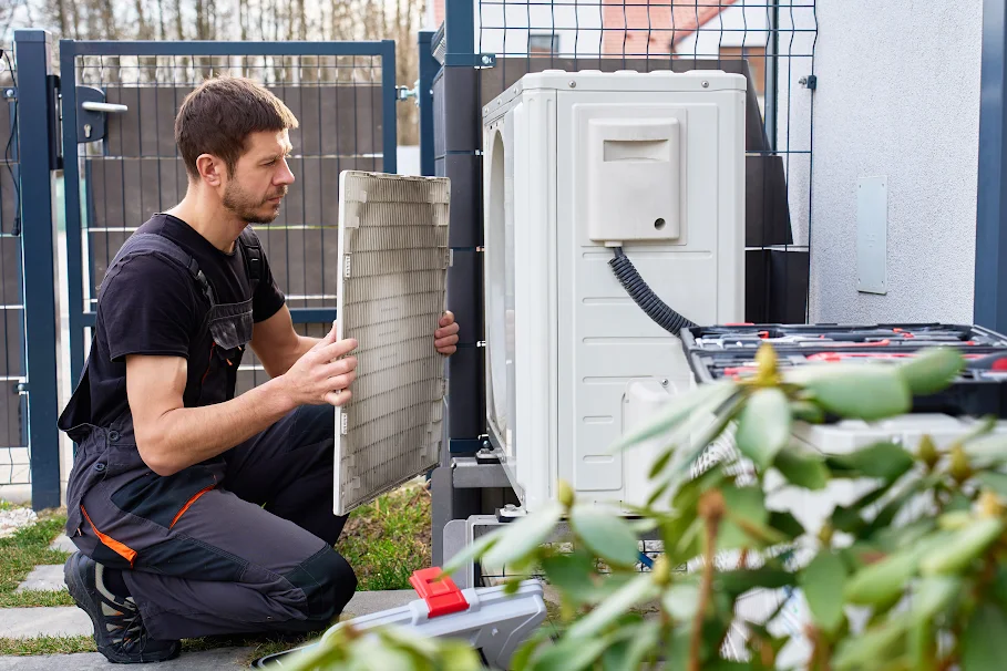 A technician inspecting an outdoor air conditioning unit, holding a filter in one hand, surrounded by greenery while checking for signs of poor indoor air quality and ensuring cleaner airflow throughout the home.