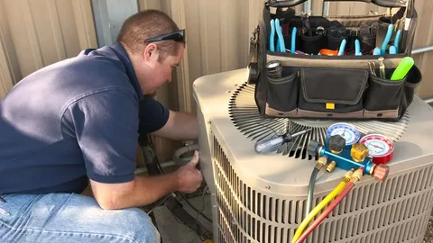 A technician in a blue shirt performs HVAC repair on an outdoor HVAC unit, with a tool bag and manifold gauge set resting on top of the beige metal cabinet.