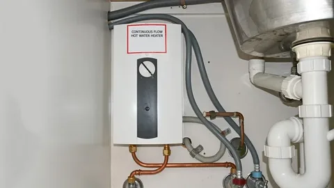A white continuous flow hot water heater mounted on a wall under a sink, connected to copper pipes, grey hoses, and white drainage pipes during a Water Heater Repair service.