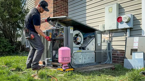 A technician in a black shirt and cap works on an open outdoor HVAC unit in Bath Township, using tools while a pink refrigerant tank sits on the grass nearby.